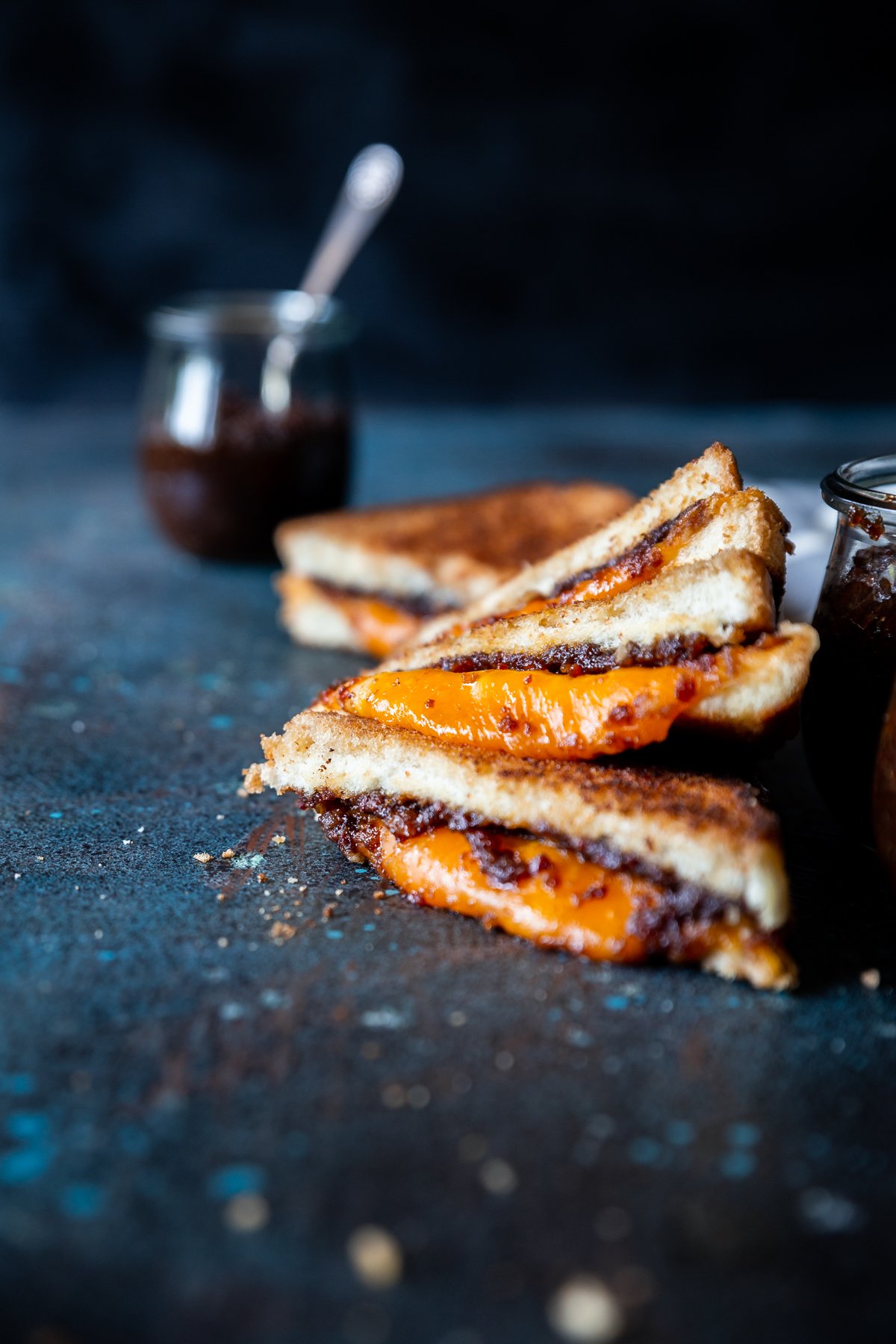 Bacon jam in small tulip jars, spoon with bacon jam on it, bacon jam grilled cheese triangles stacked on each other