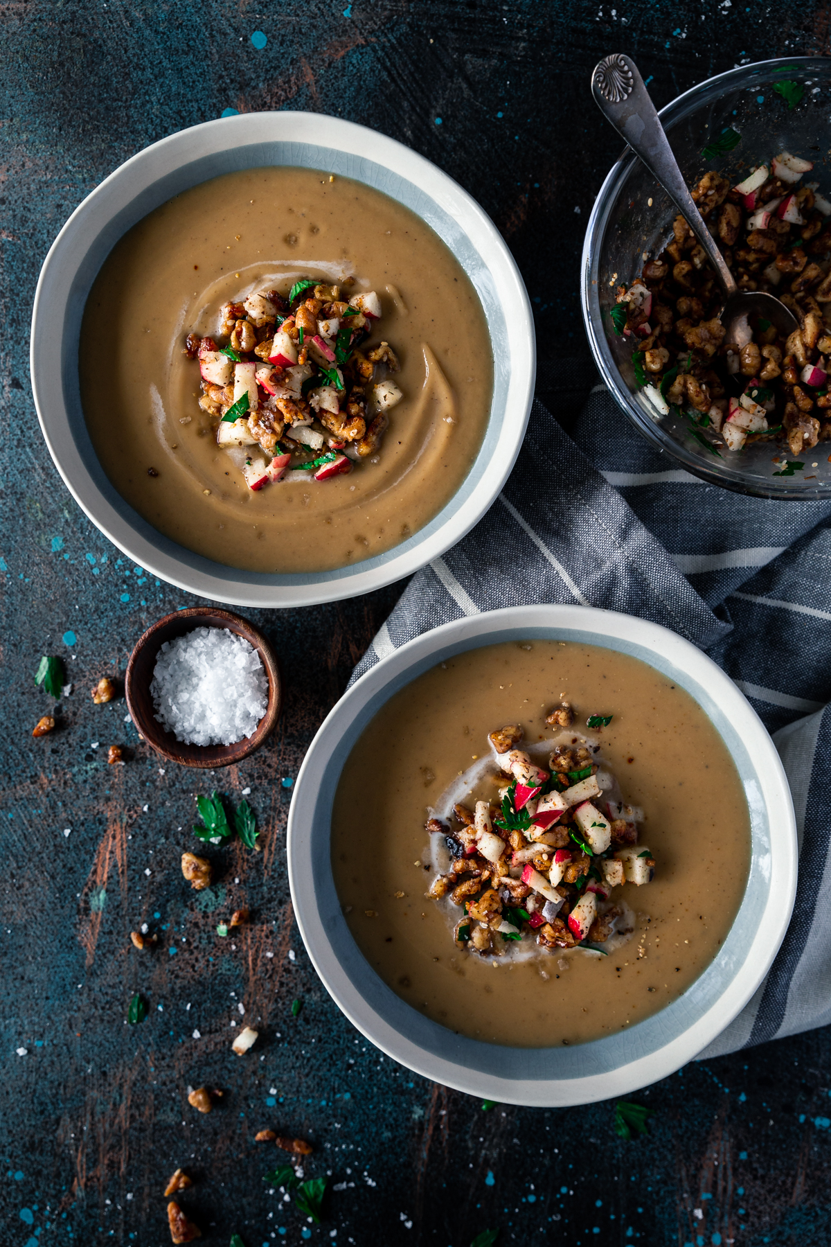 golden brown creamy parsnip soup in light dusty blue bowls with white rims, with walnut and diced apple topping in the middle. a small bowl of topping sits to the top right, and a small wooden dish with salt sits center-left.