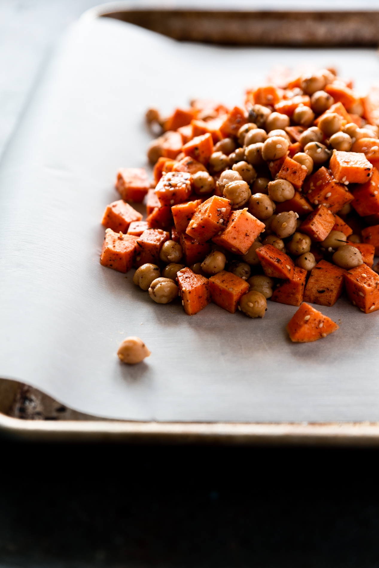 forty-five degree angled shot of a baking sheet lined with parchment paper containing uncooked diced sweet potato and chickpeas coated in olive oil, za'atar seasoning, and salt.