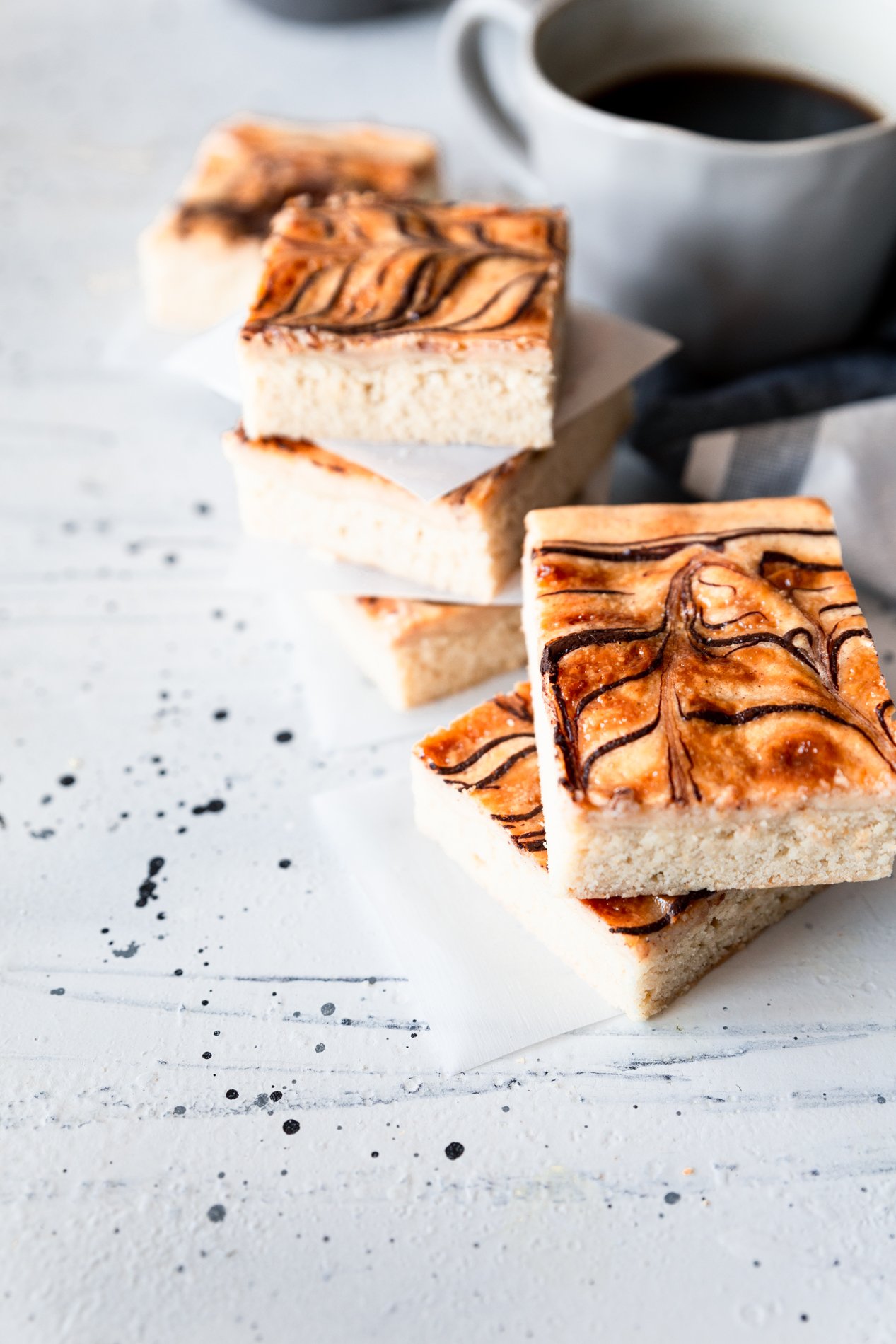 forty-five degree angled view of churro cheesecake cookie bars with dulce de leche and dark chocolate ribbons. Slices are staked on top of each other on a white background, with a cup of coffee to the upper right of the image on a blue cloth.