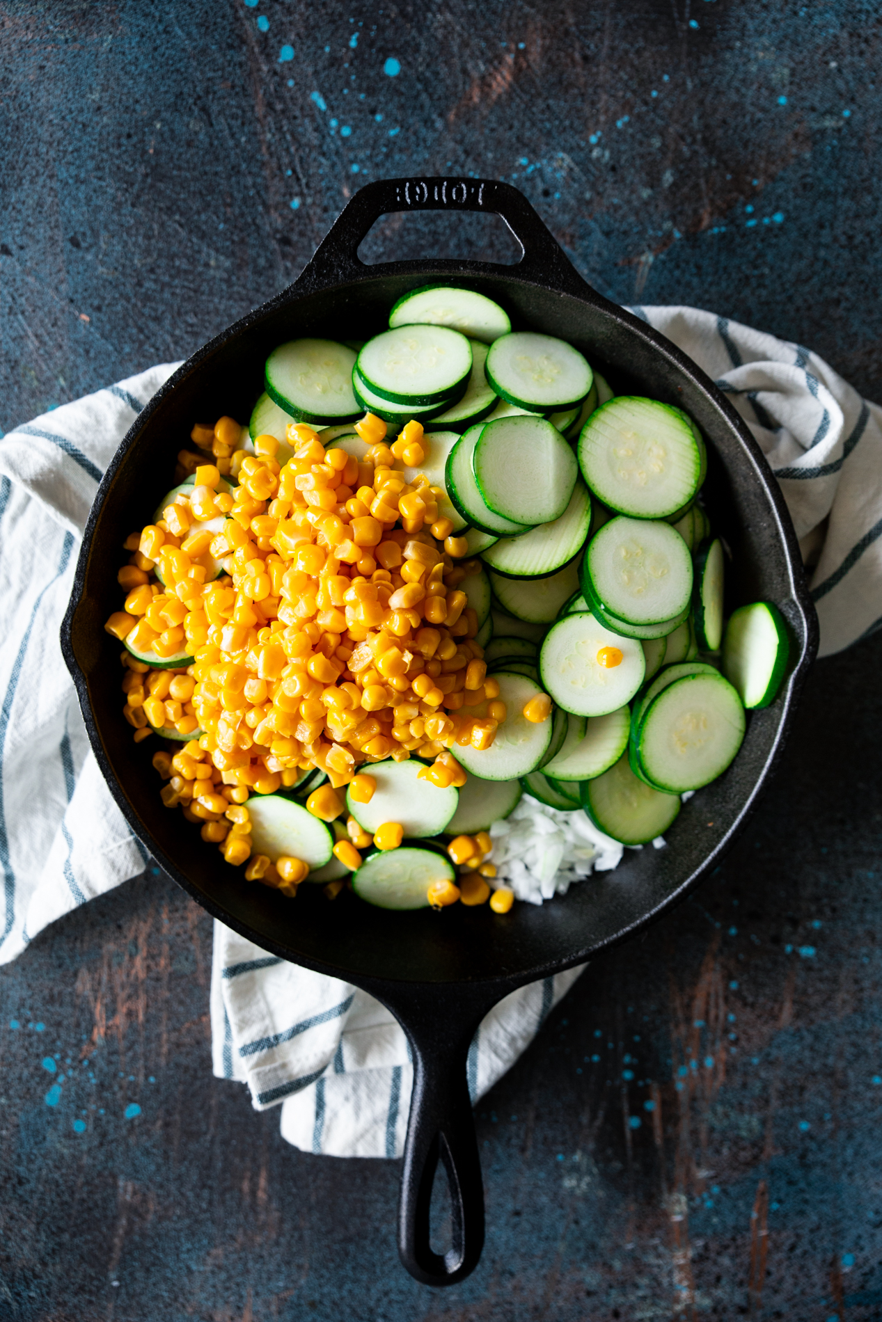 overhead view of a cast iron skillet filled with uncooked onion, zucchini, and corn. the skillet is on top of a striped cloth.
