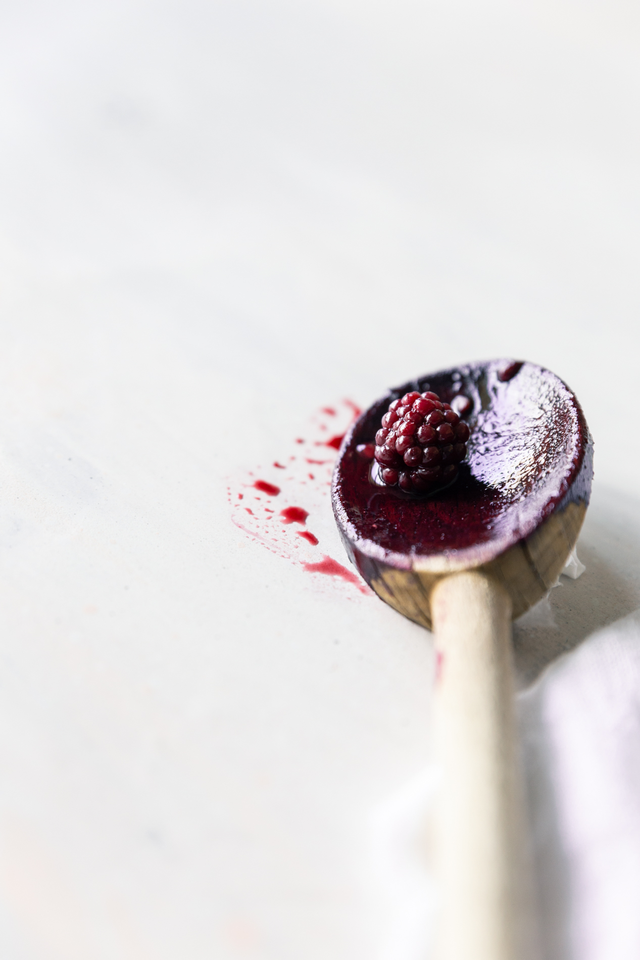 forty-five degree angled view of a wooden spoon with blackberry juices on it and a single small blackberry.
