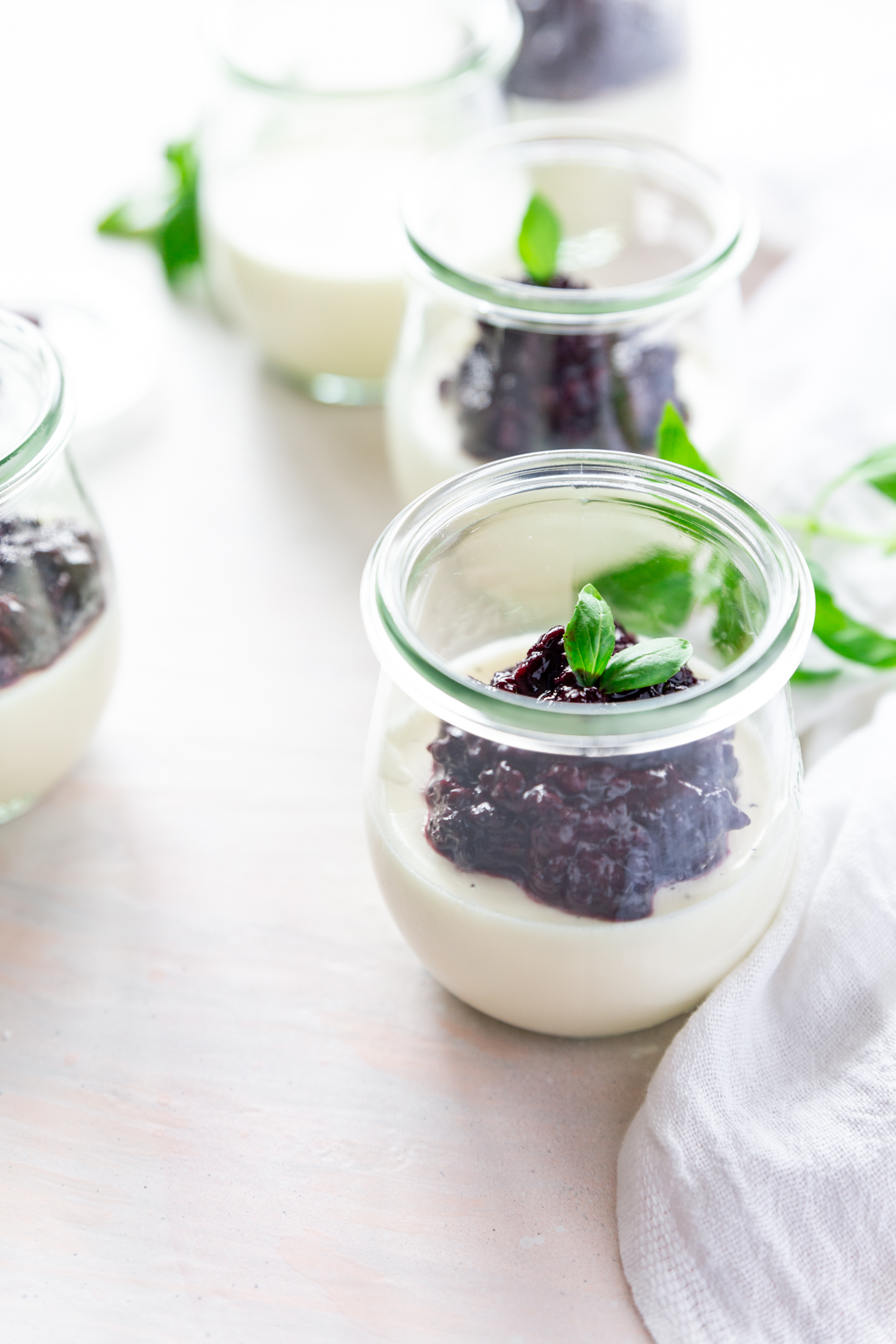 forty-five degree angled shot of jars of vanilla bean panna cotta with basil blackberry compote, surrounded by fresh basil leaves and pleasantly backlit.