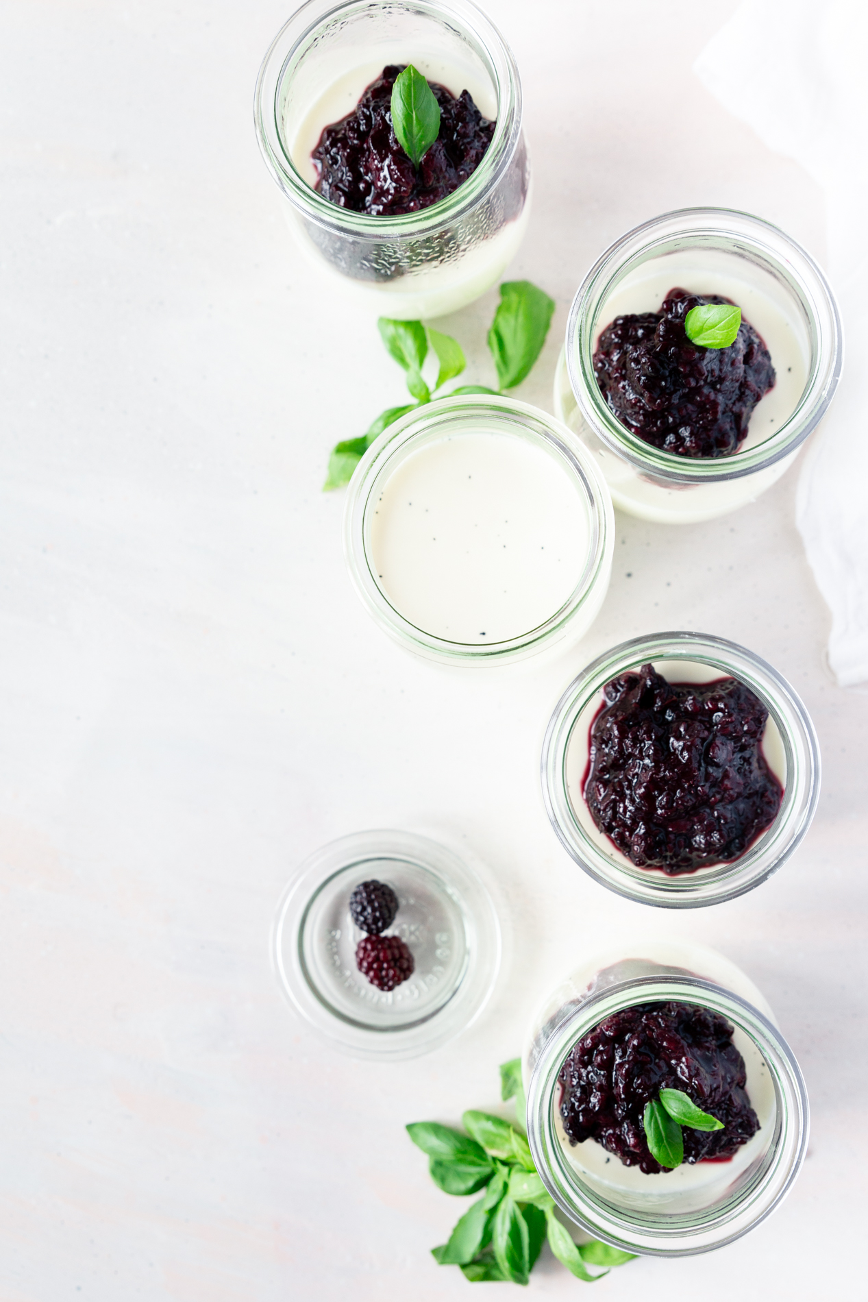 overhead view of jars of vanilla bean panna cotta with basil blackberry compote, surrounded by fresh basil leaves.