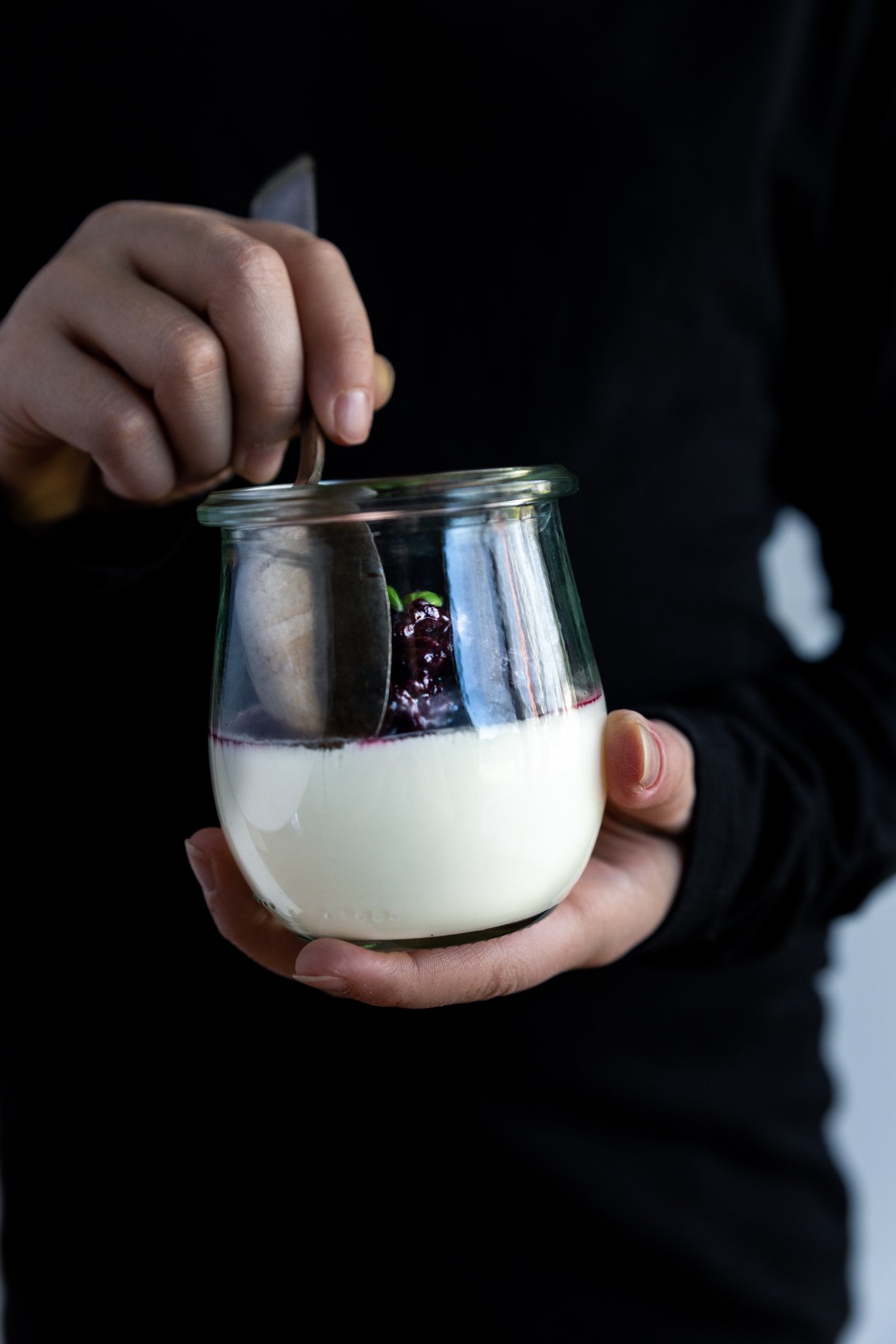straight-ahead view of a person in a black shirt holding a jar of vanilla bean panna cotta with basil blackberry compote. the right hand is holding a spoon and in the middle of getting a scoop of dessert.