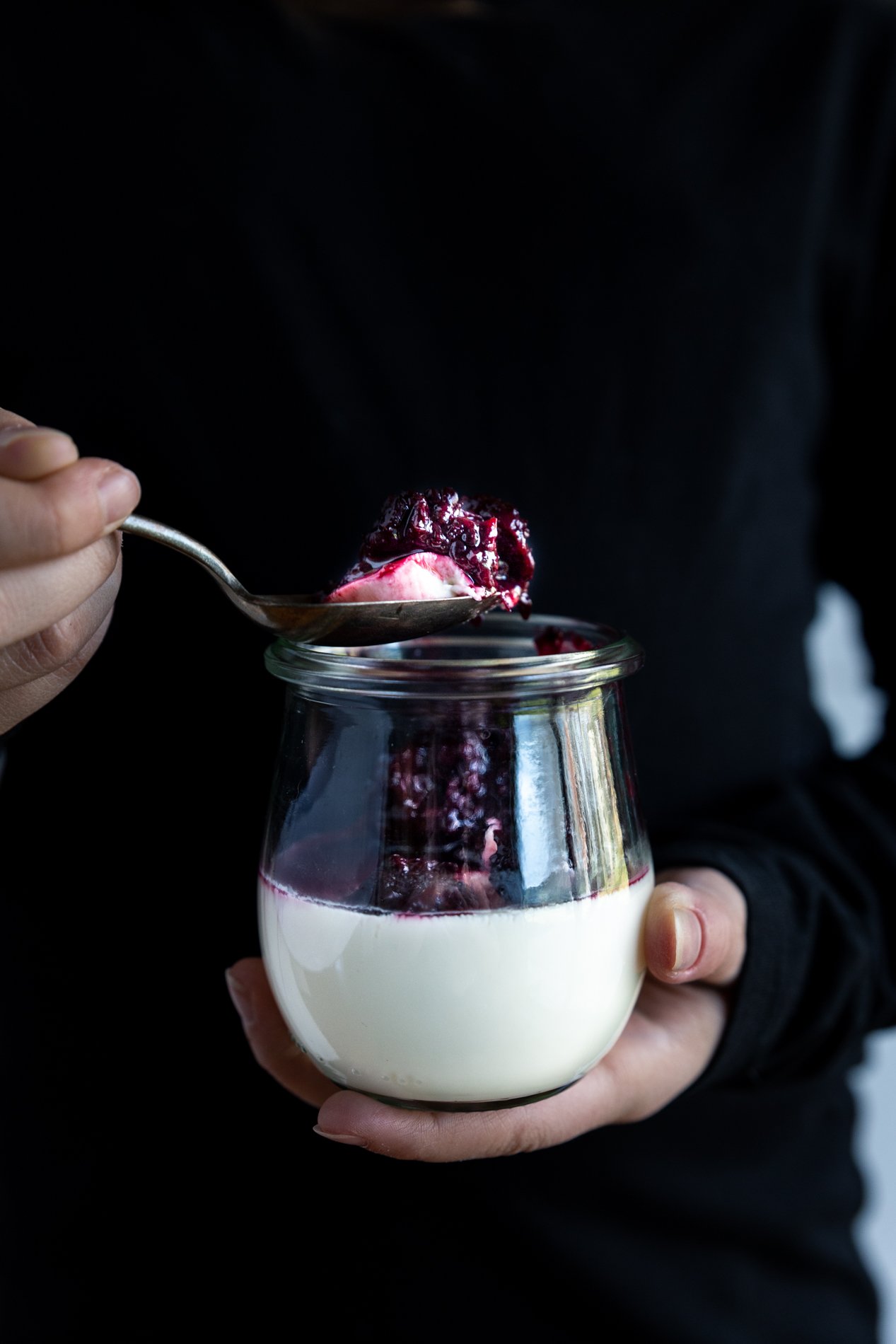 Straight ahead view of a person holding a jar of vanilla bean panna cotta with basil blackberry compote. In right hand is a spoon with a scoop of the dessert, person is wearing a black shirt.