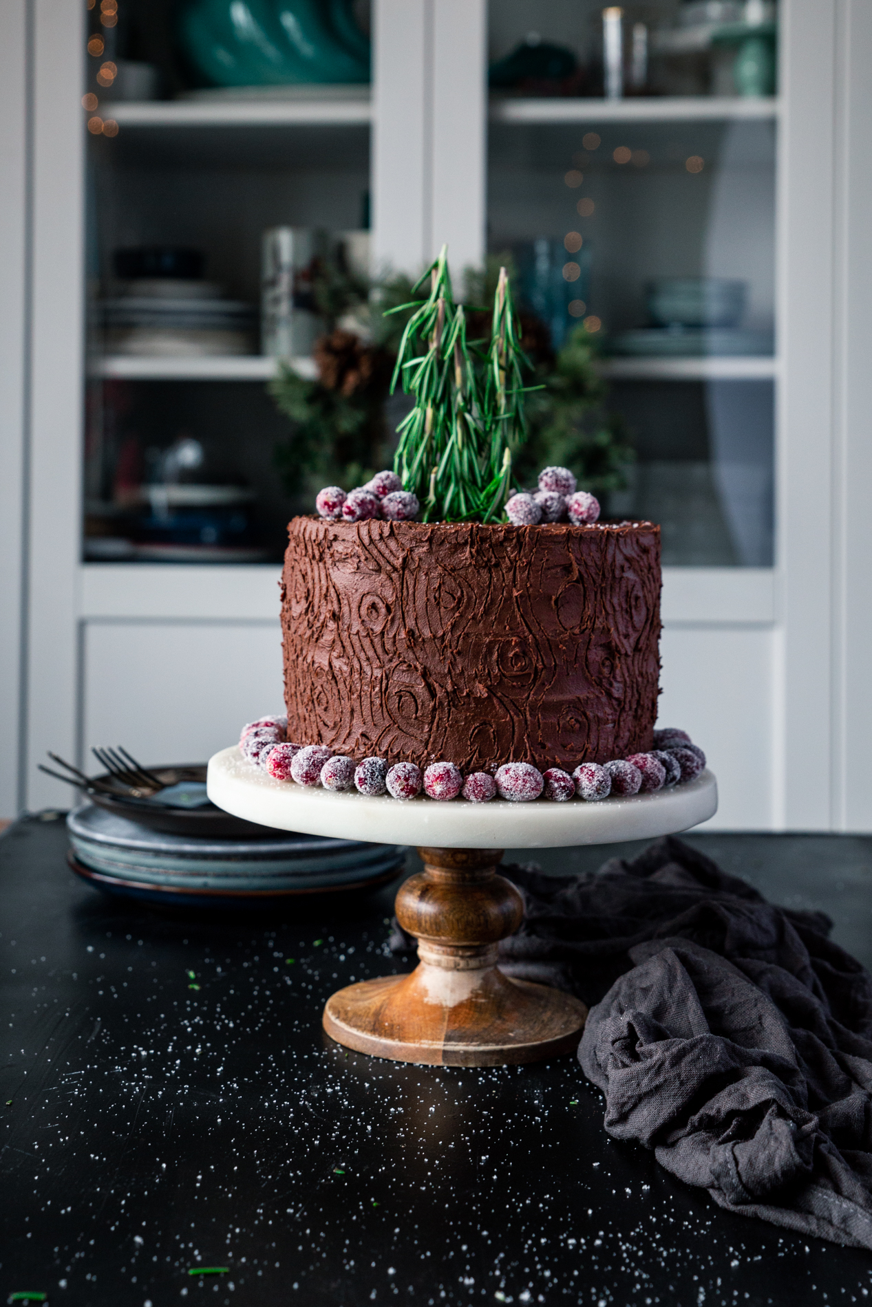 straight-forward view of a chocolate cake on a cake stand with a tree stump design, surrounded by sugared cranberries, with sugared cranberries on top and rosemary sprigs acting as trees.