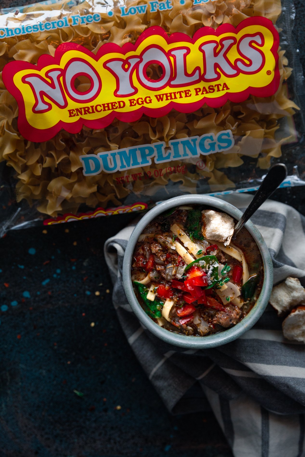 overhead view of a bowl of easy sausage and bell pepper noodle soup on top of a gray striped napkin, with a bag of No Yolks Dumplings noodles directly above it.