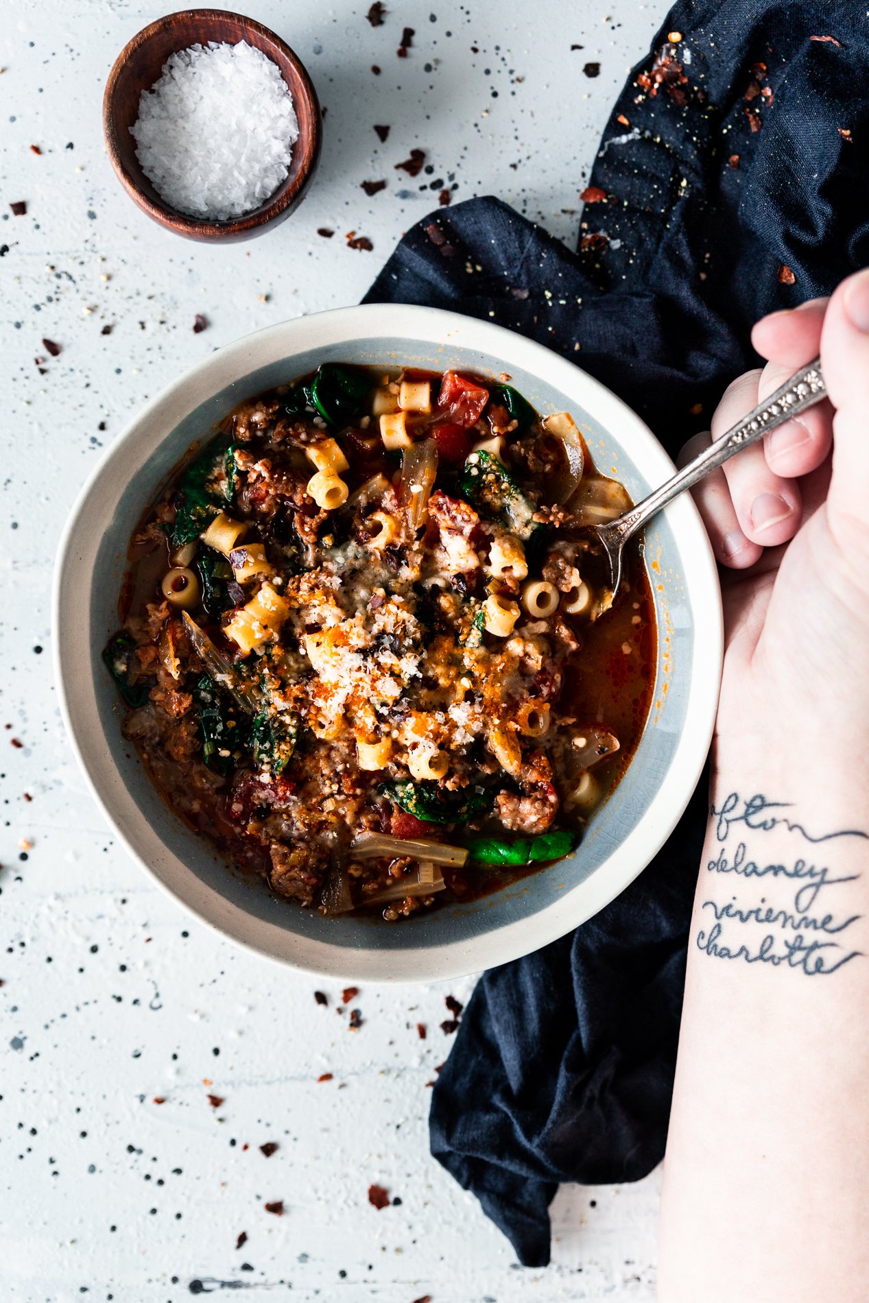 overhead view of a bowl of italian sausage soup with spinach, pasta, and caramelized fennel from minnesota food blogger a simple pantry. a small bowl of garnishing salt is toward the top of the image and an arm holds a spoon inside the bowl of soup.
