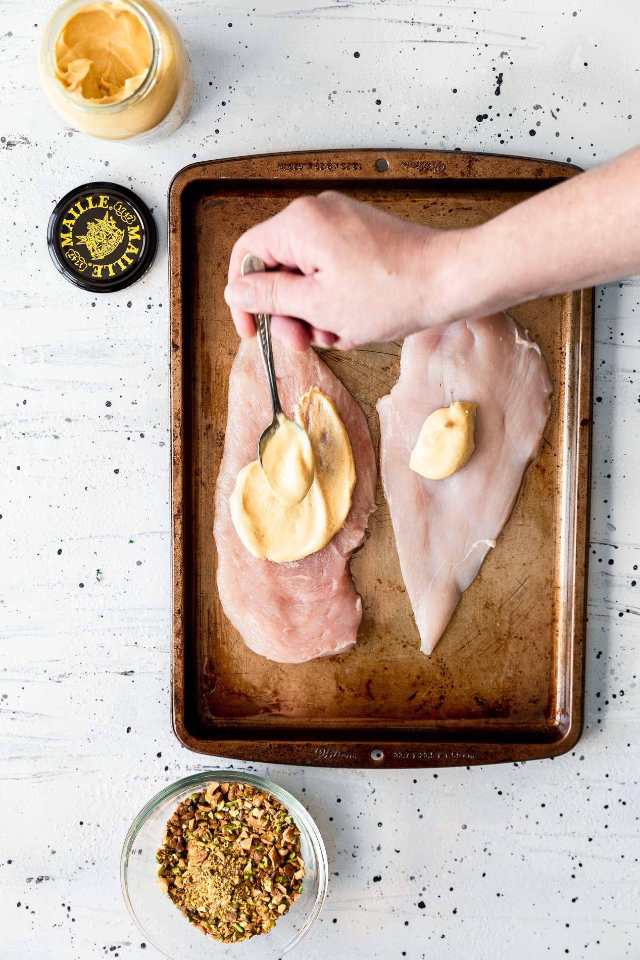 overhead view of dijon mustard being spread on chicken breasts before coating with pistachios in a small bowl at the bottom of the frame.
