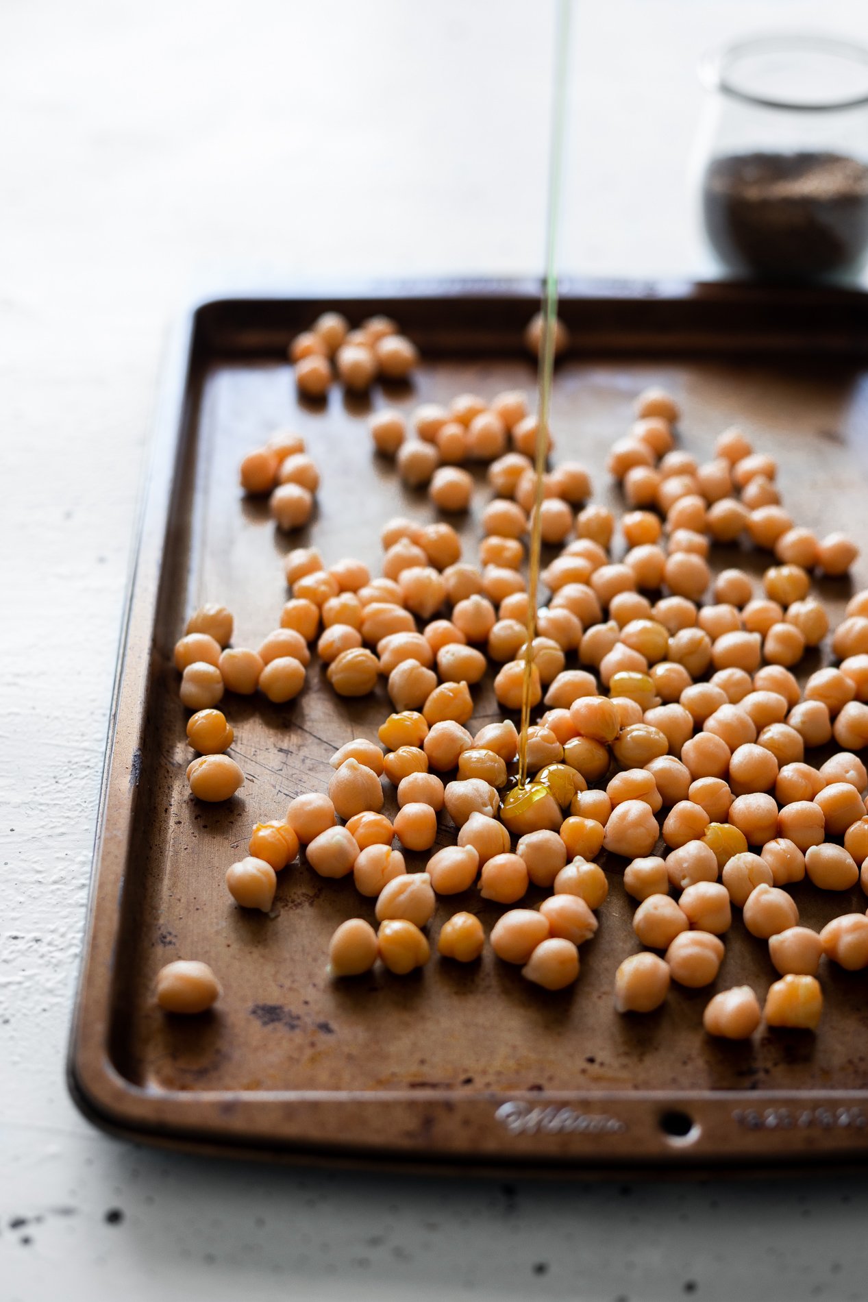 side view of chickpeas on a baking sheet with olive oil being drizzled over them, from minnesota food blogger asimplepantry