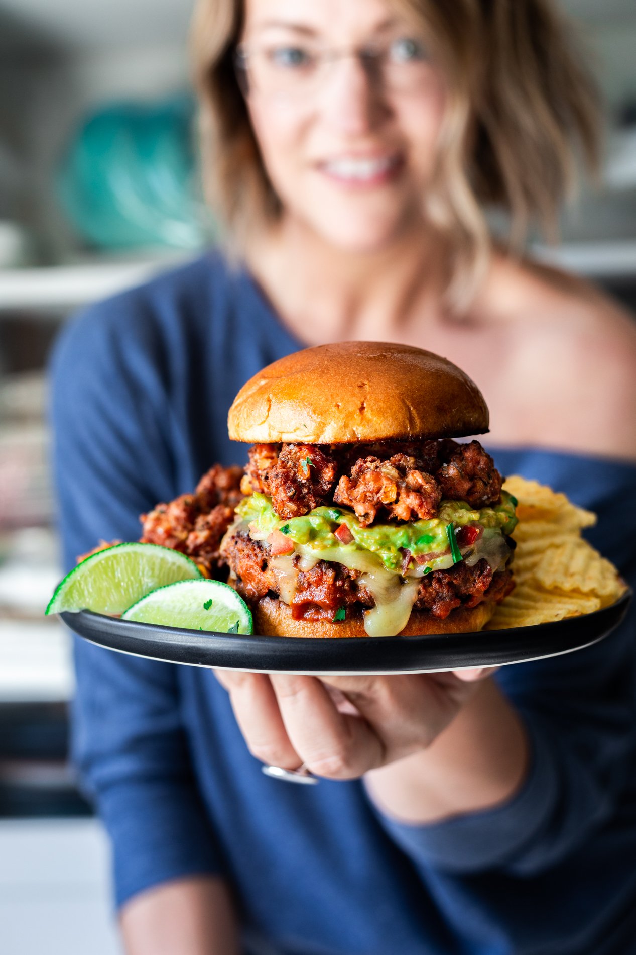 straight-ahead view of chorizo burger with mexican street corn fritters on a plate with limes and ruffled chips, held by minnesota food blogger karly gomez