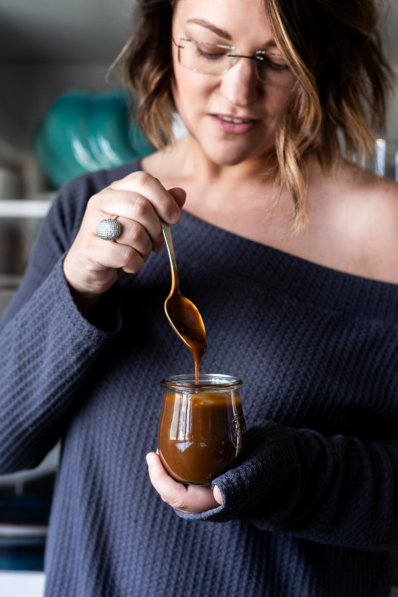 straight ahead view of karly gomez holding a jar of vegan caramel sauce with a spoon drizzling sauce back into the jar