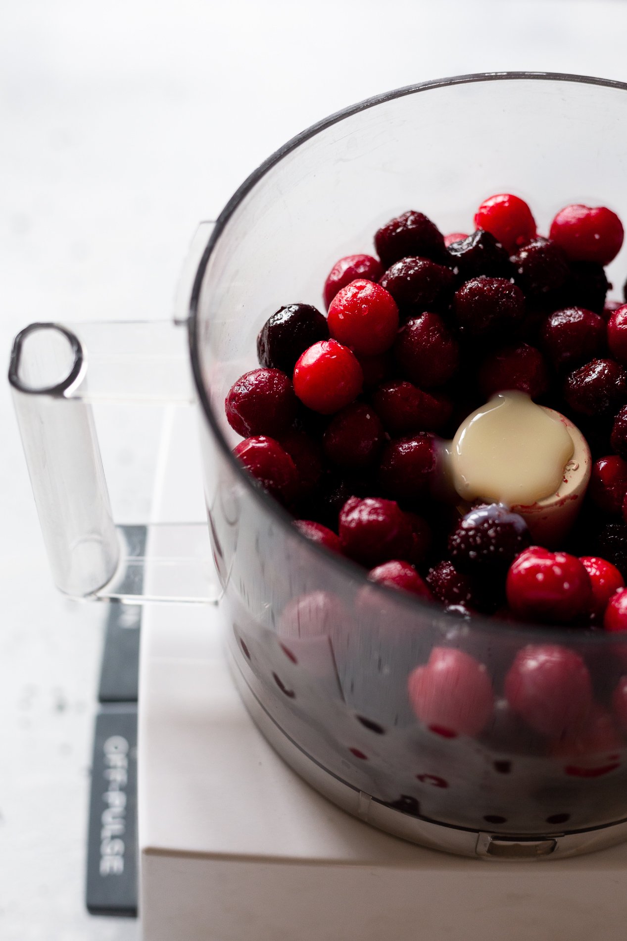 angled view of a food processor bowl filled with frozen cherries, vanilla, sweetened condensed milk, and a sprinkle of salt