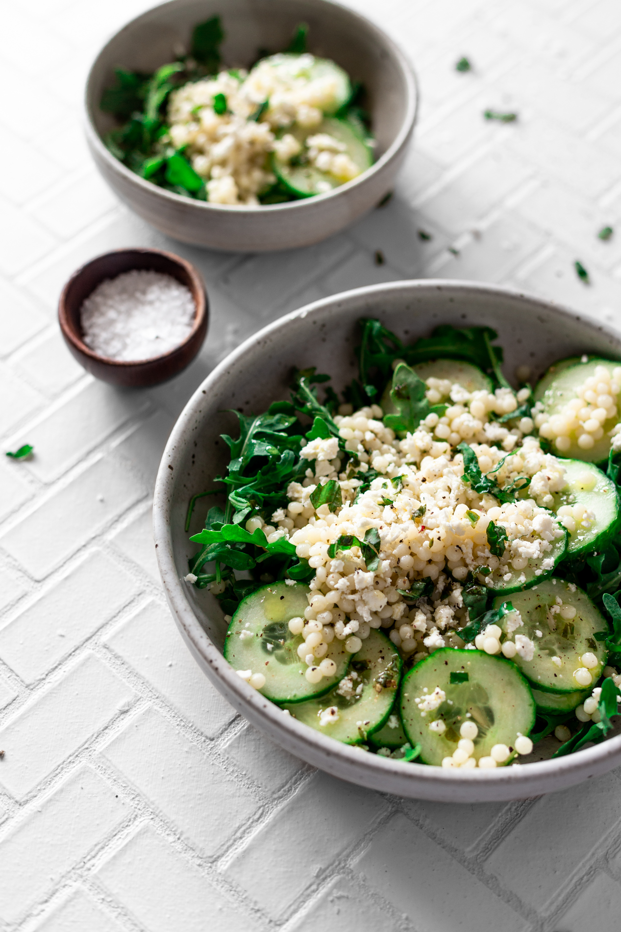 angled view of two bowls of cucumber salad recipe with lemon basil vinaigrette with a small bowl of flake sea salt