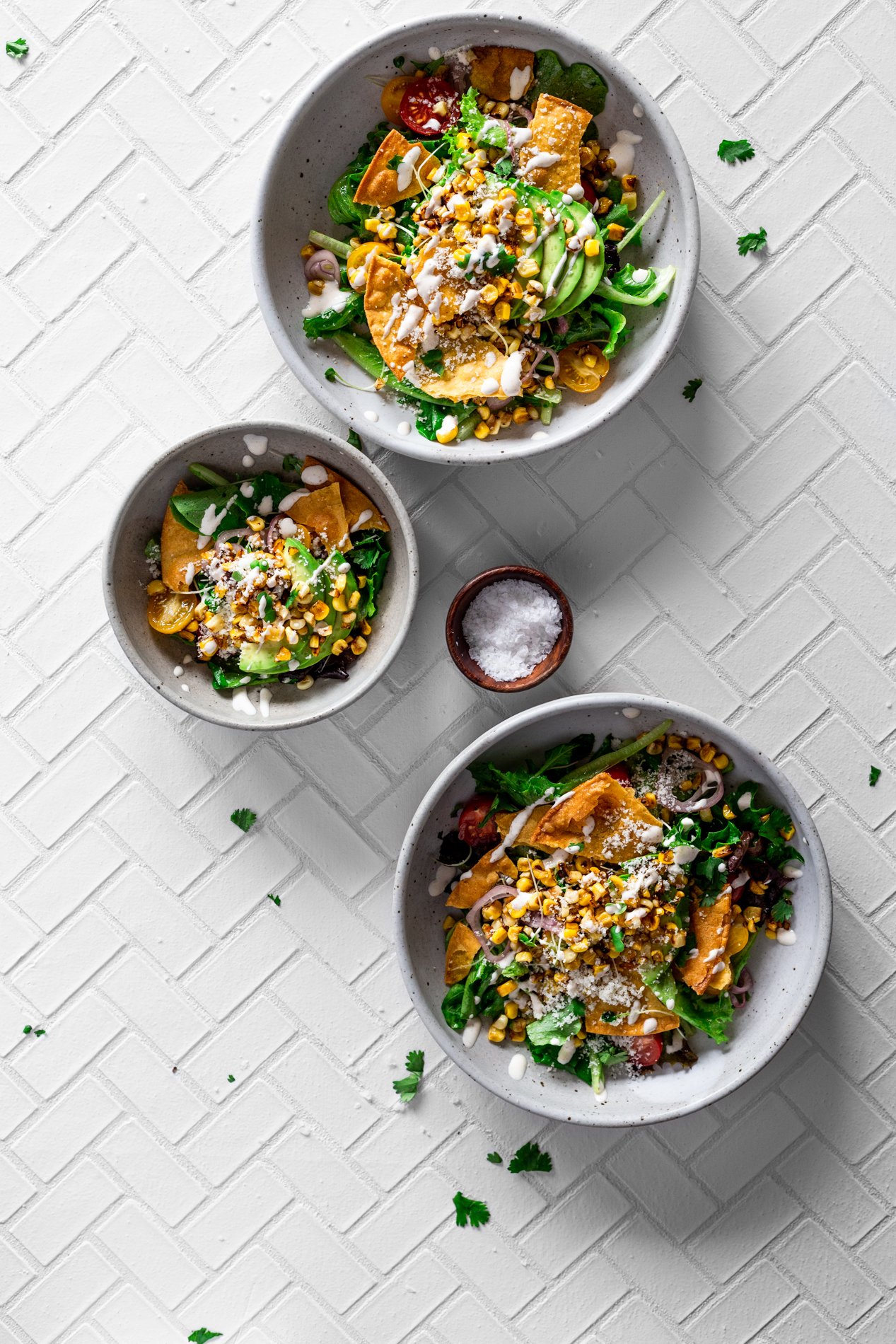 overhead view of two large and one small bowl of mexican street corn salad with a tiny bowl of salt beside them, cilantro sprinkled around the bowls