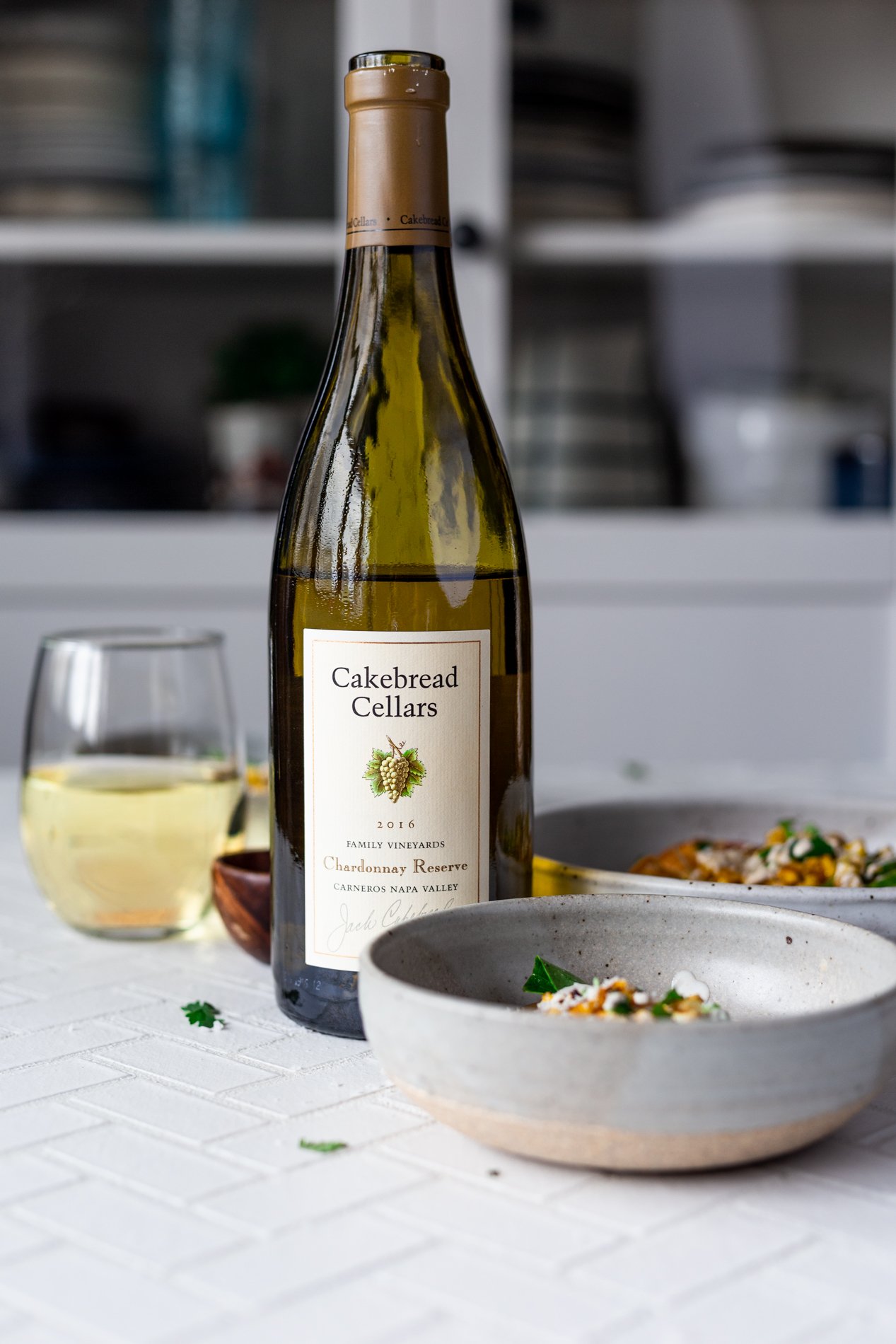 straight-ahead view of a bottle of Cakebread Cellars Chardonnay Reserve surrounded by bowls of mexican street corn salad and a glass of wine