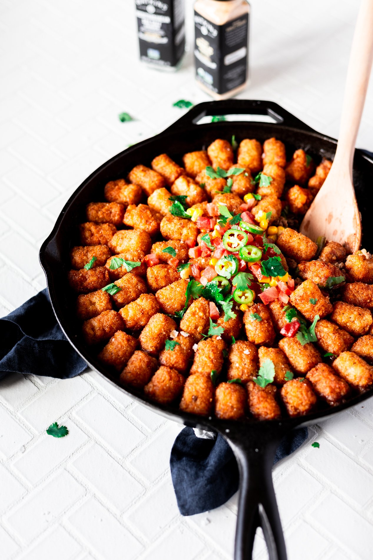 angled view of a skillet full of not yo mama's tater tot hotdish with seasoned tots and seasoning bottle towards the top of the image. a spoon has taken a scoop of the hotdish