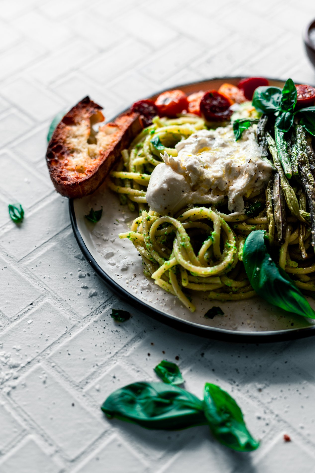angled view of a plate of walnut pesto pasta with roasted summer vegetables and a dollop of burrata and crusty toasted bread