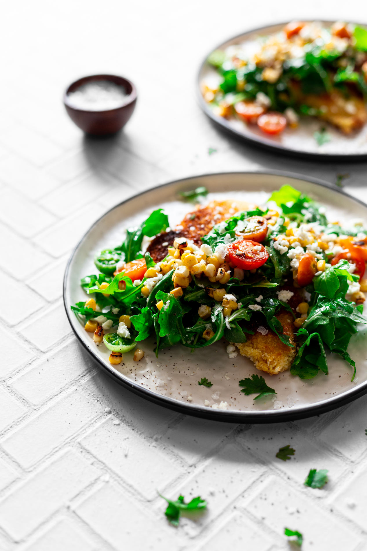 angled view of two plates of easy chicken milanese recipe with charred corn, tomato, and arugula salad with a small dish of flake salt in the background
