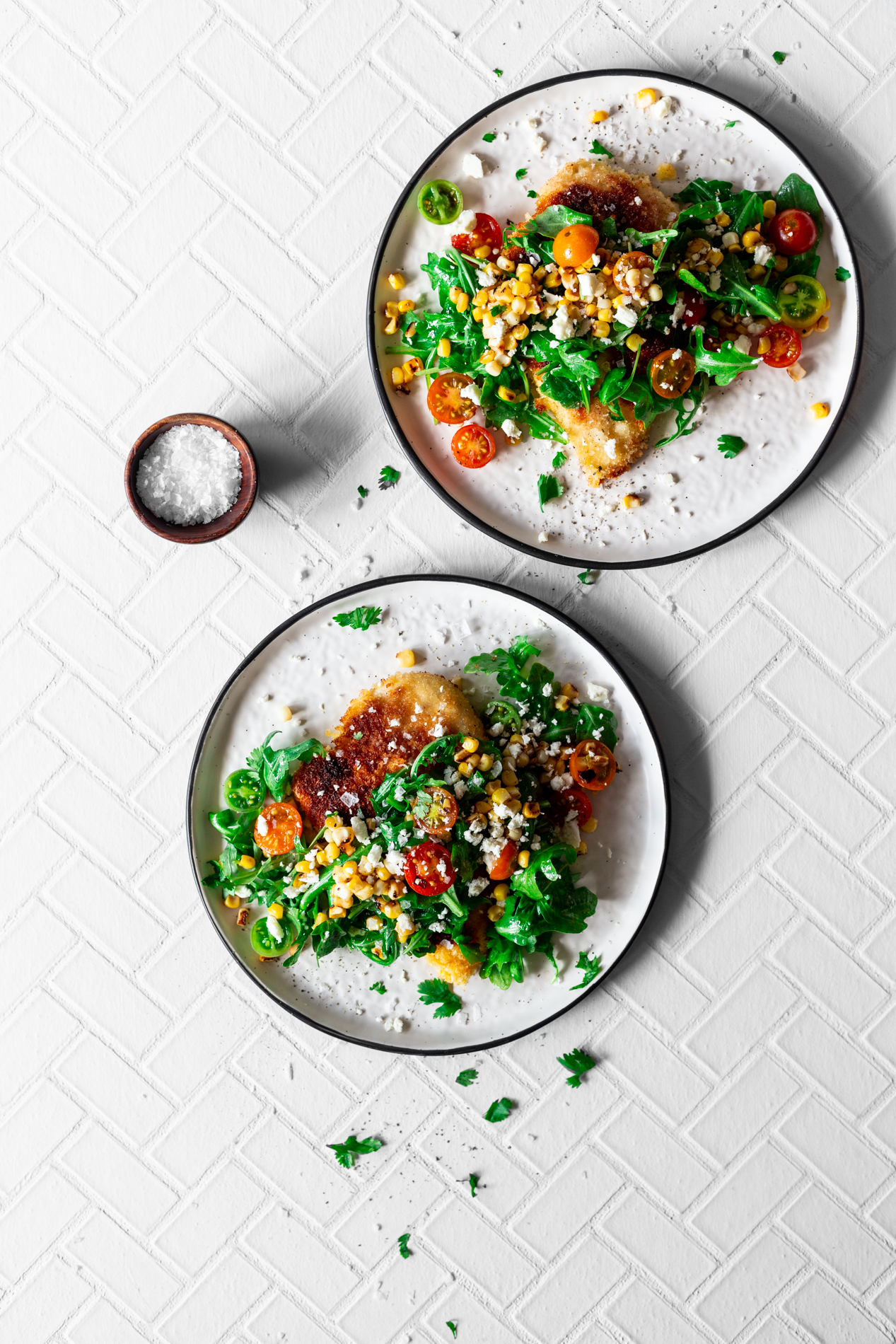 overhead view of two plates of easy chicken milanese recipe with charred corn, tomato, and arugula salad with a small bowl of flake salt to the side