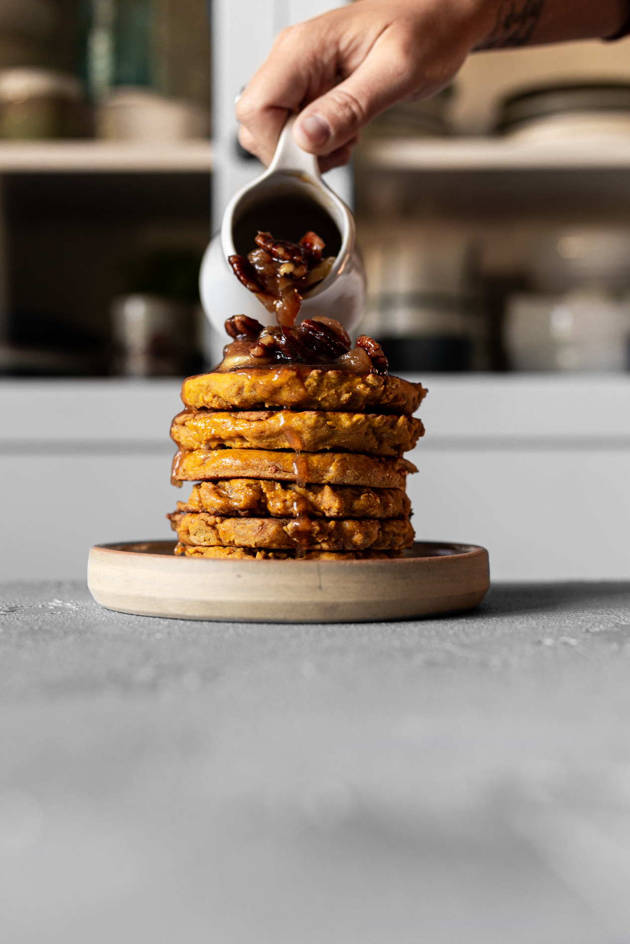 hand pouring caramelized bananas and pecans on a stack of pumpkin bread griddle cakes