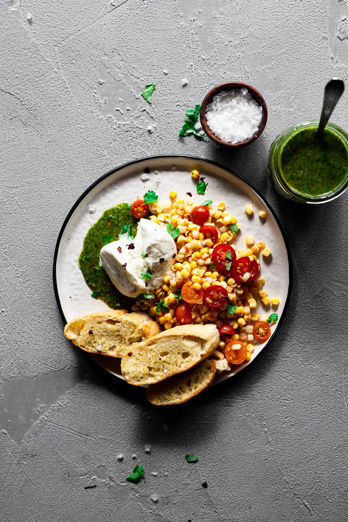 a plate with chimichurri topped with burrata and surrounded with tomato and corn salad
