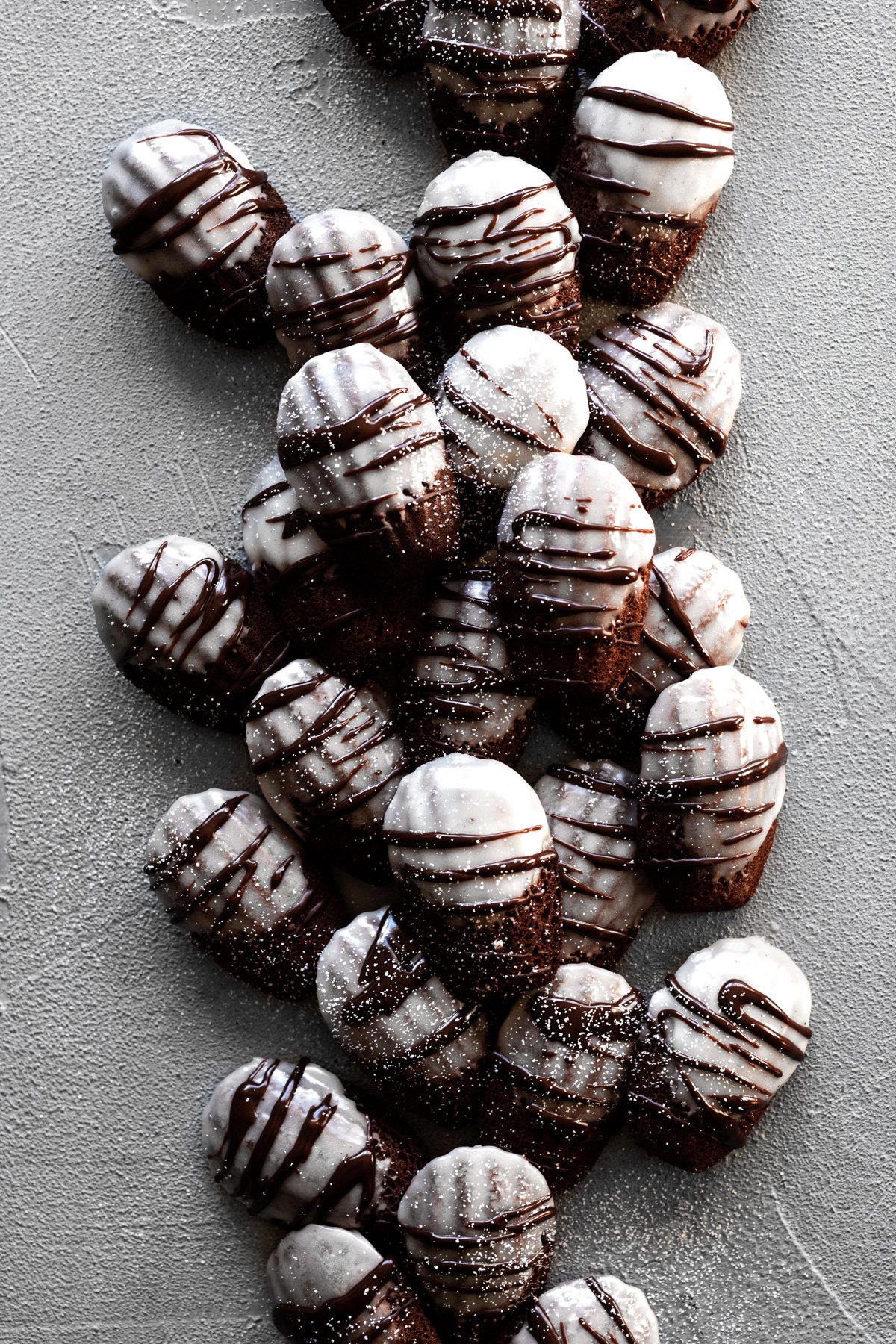 pile of hot chocolate madeleine cookies with marshmallow frosting, chocolate drizzle, and powdered sugar